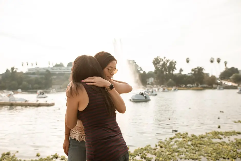 Two women embracing in a park by a lake with a fountain, captured during a sunny day.