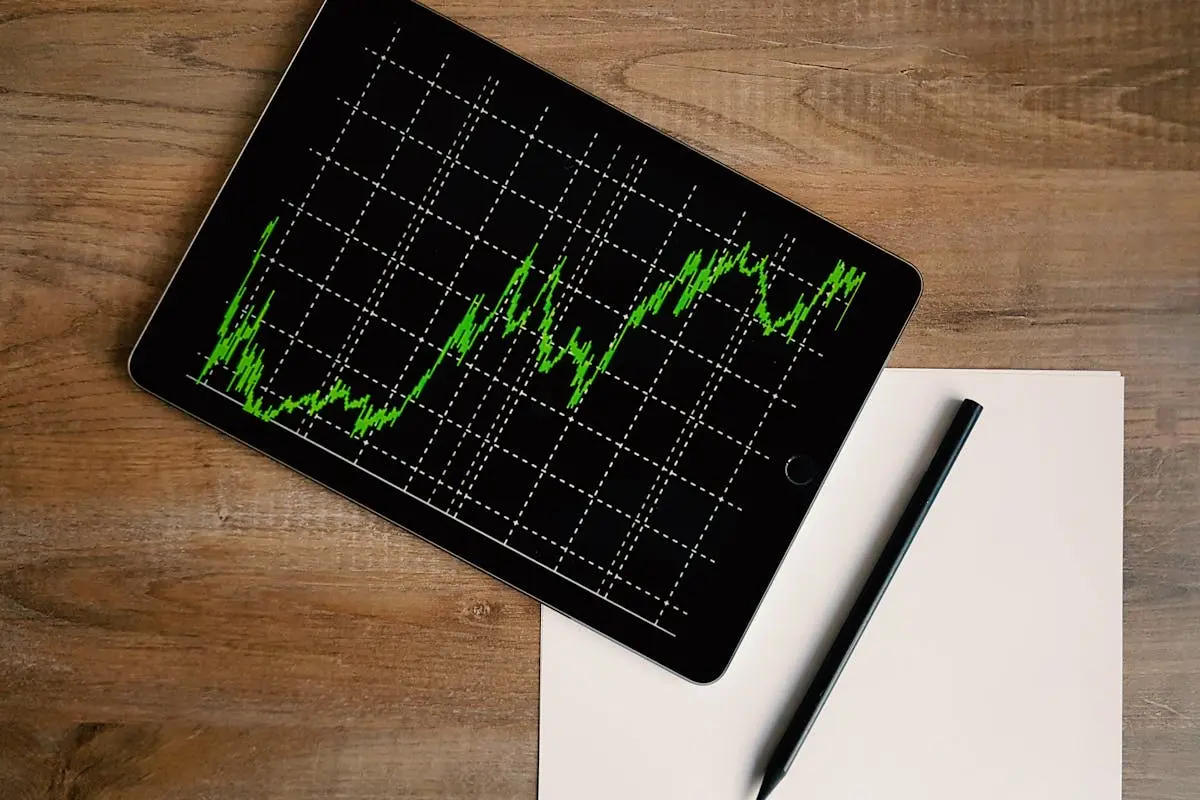 Flatlay of an outsourced cfo ipad displaying stock market graph on a wooden desk with a pencil and paper.
