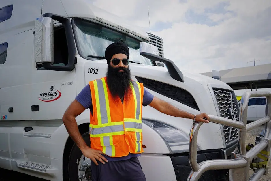 Sikh truck driver with turban and safety vest standing beside a white truck on a sunny day.