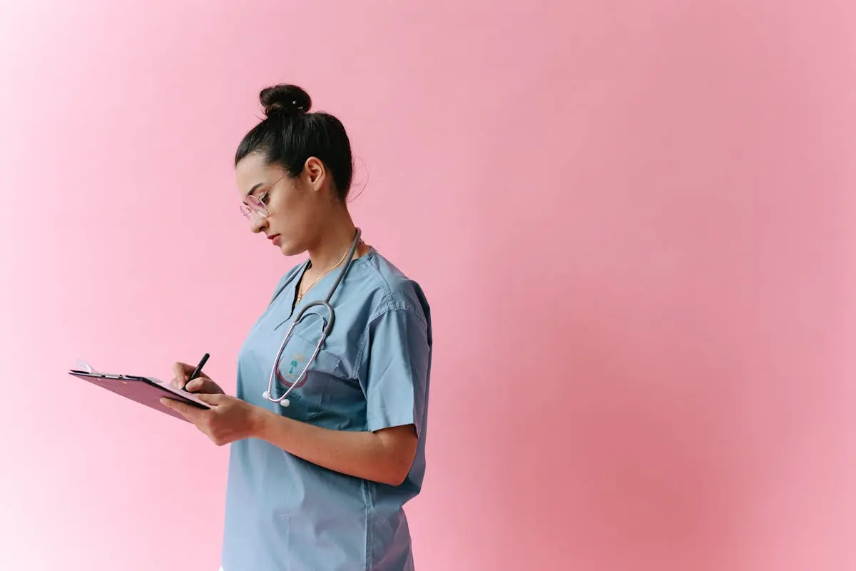 Female nurse in scrubs writing on a clipboard with a stethoscope on a pink background.