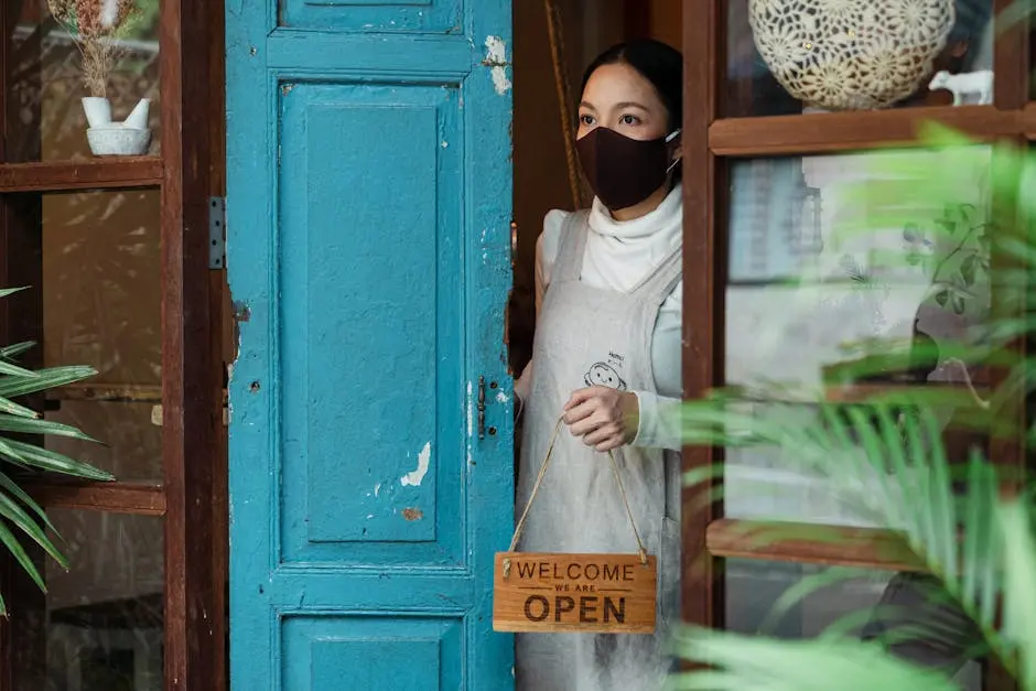 Calm young Asian female wearing casual clothes and face mask standing at shabby rural shop doorway and removing open sing