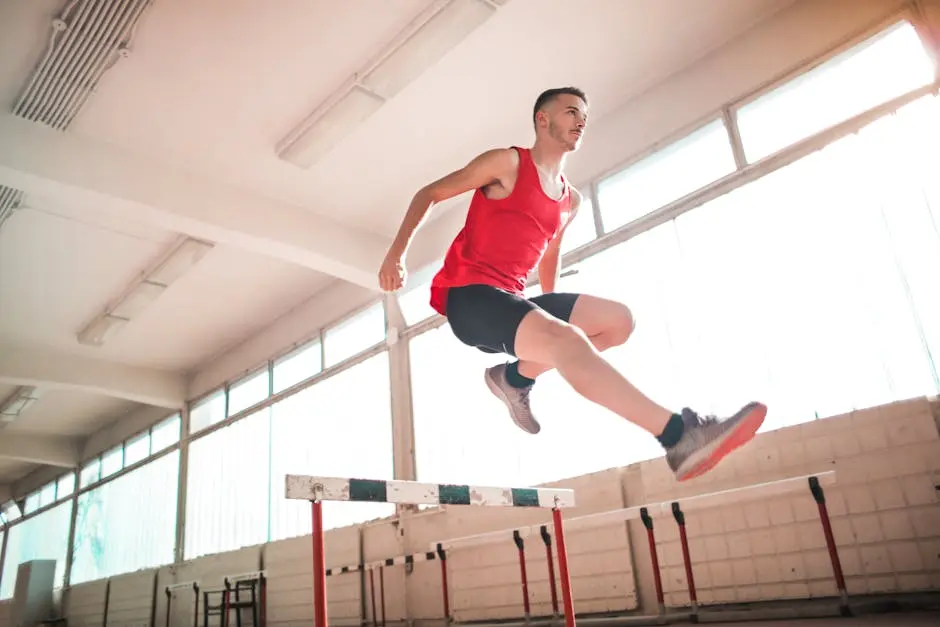 A young male athlete in a red outfit jumps over a hurdle indoors, showcasing agility and determination.