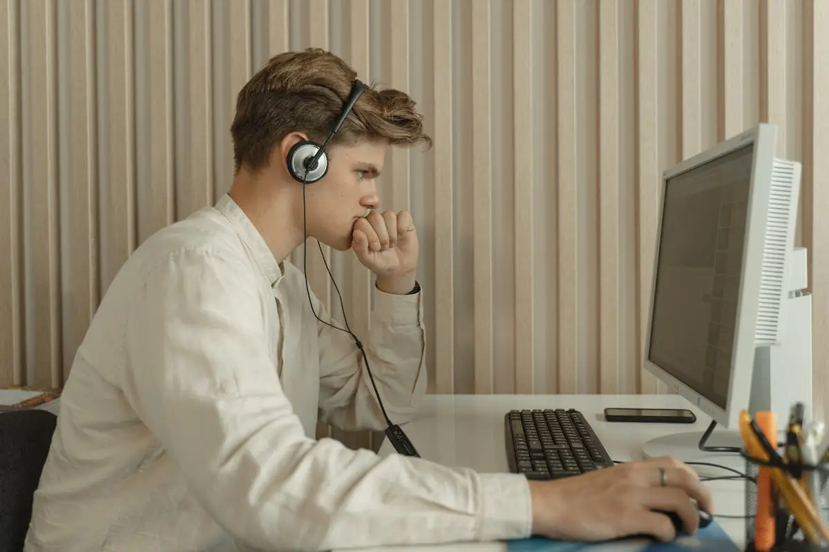 A young man wearing headphones is intently working on a desktop computer in an office setting.