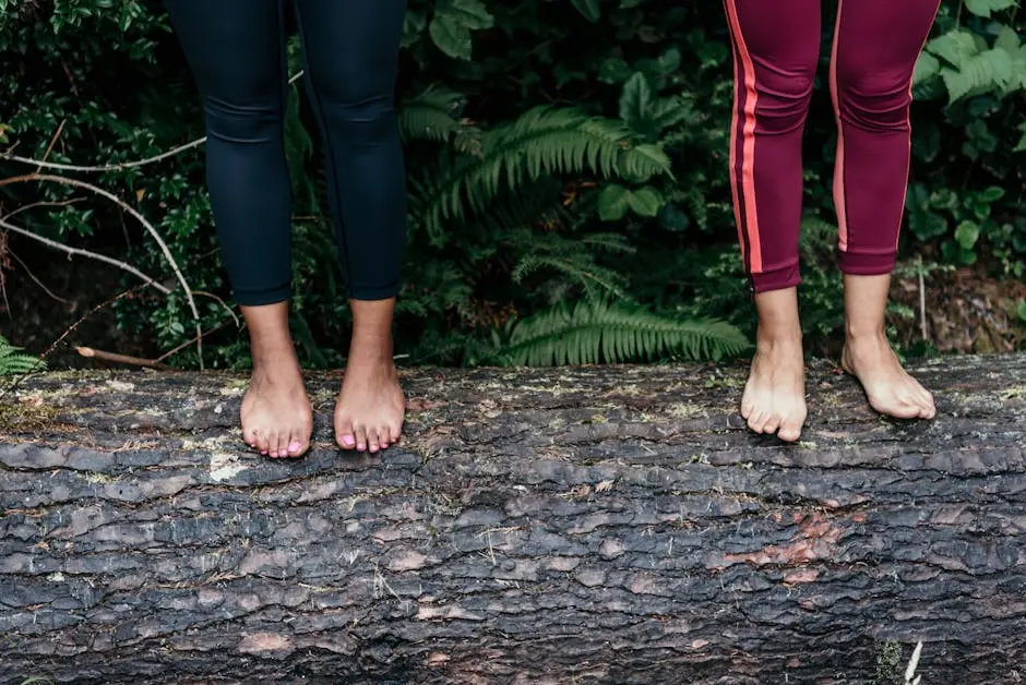 Two women standing barefoot on a log amidst lush greenery in a forest setting.