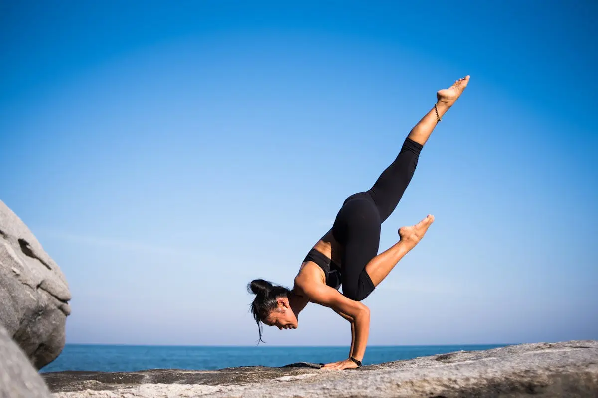 Woman performing a yoga pose on a rocky beach with ocean view and clear blue sky.