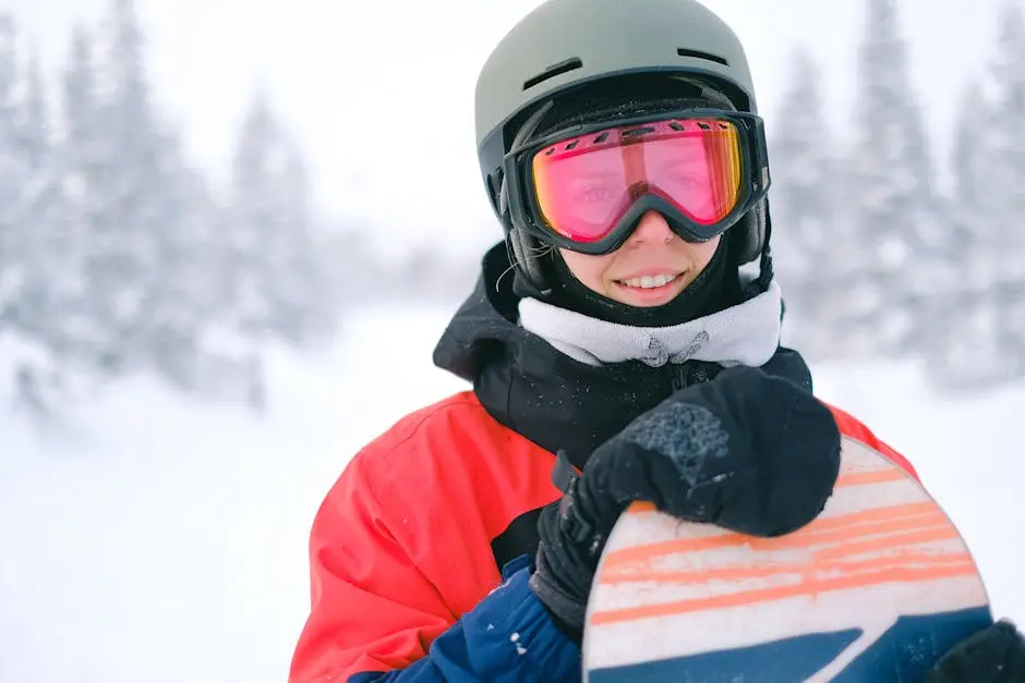A snowboarder in winter gear smiling while holding a snowboard in snowy landscape.