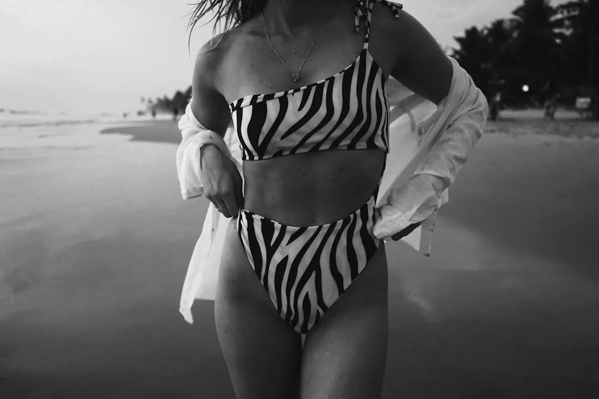 Woman in zebra print swimsuit walks on beach at sunset, showcasing summer fashion in a black and white setting.