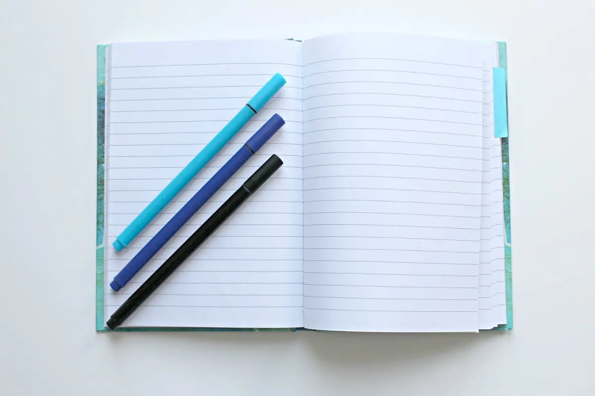 Open lined notebook with colorful pens on a white surface, perfect for note-taking and office tasks.