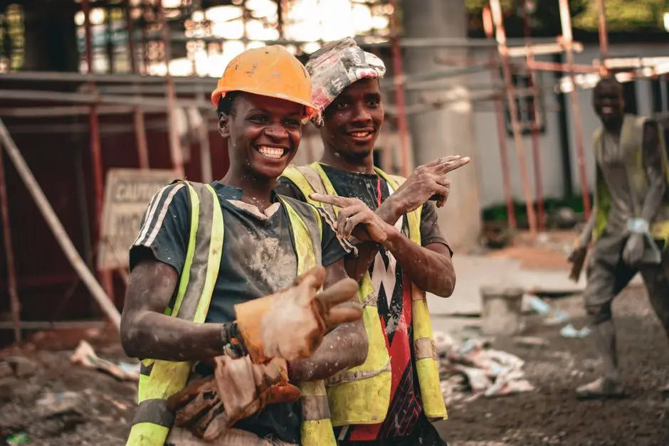 Two smiling construction workers in hard hats and safety vests pointing with joy on a construction site.