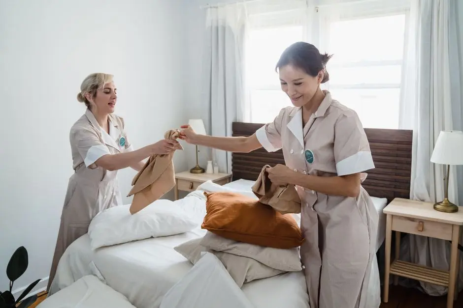 Two female housekeepers in uniform efficiently prepare a bed in a bright, well-lit room.
