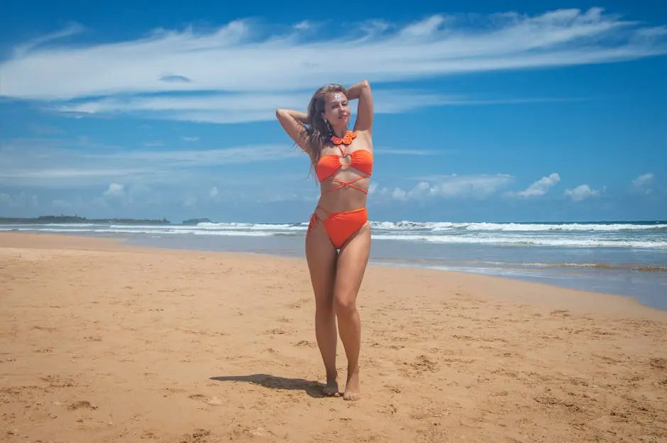 A woman in an orange swimsuit poses confidently on a sunny beach with blue skies.