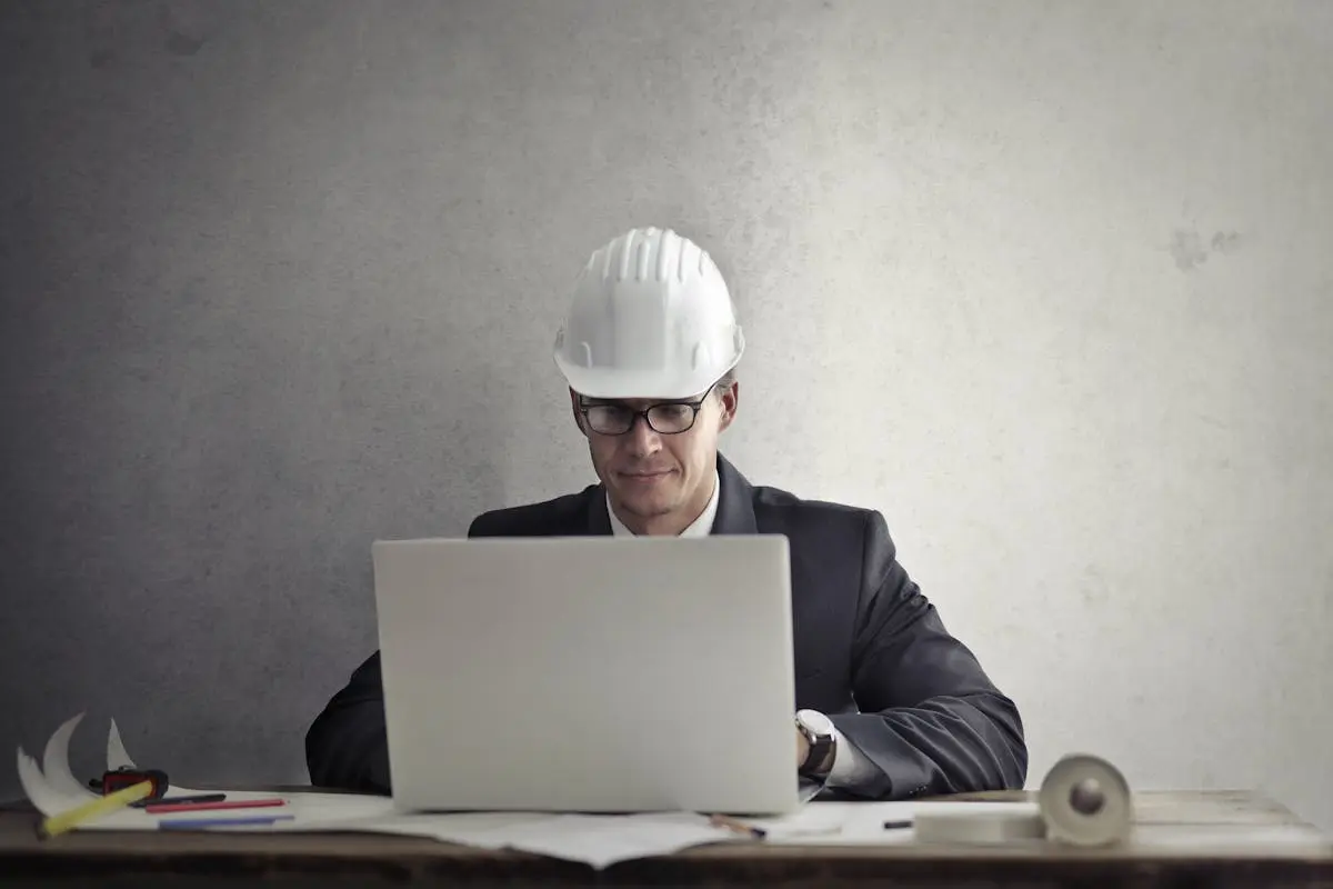 Professional business coach wearing hardhat working on construction plans at desk with laptop in office.