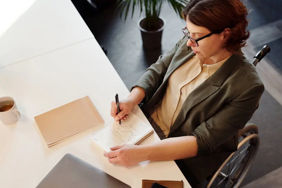 A professional woman in a wheelchair writes notes at a modern office desk, showcasing workplace diversity.