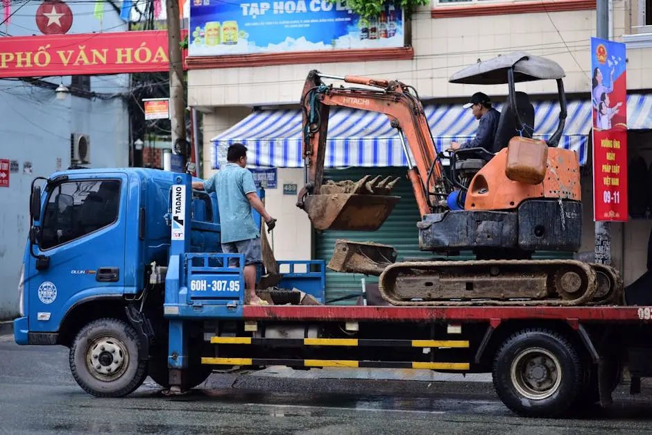 Two men and excavator being transported by truck on rainy urban street.