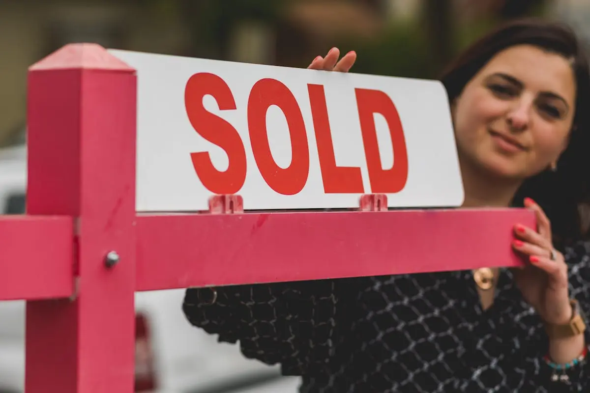 Woman smiling behind a &lsquo;Sold&rsquo; sign, symbolizing successful home purchase.