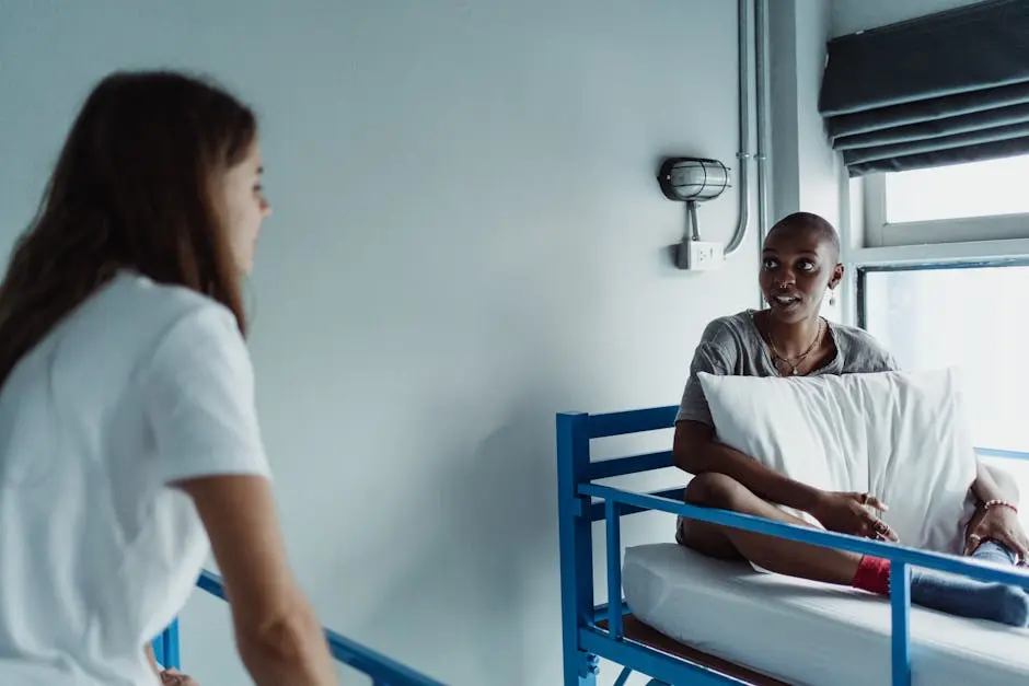 Two women talking in a hospital room, one sitting on a bed holding a pillow.