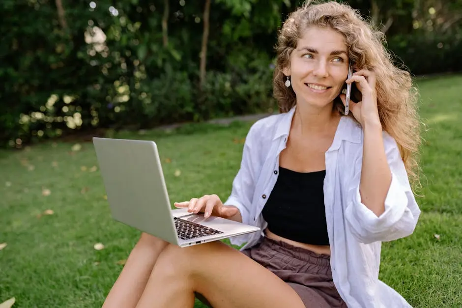 Woman sitting on grass using a laptop and talking on phone, enjoying outdoor work.