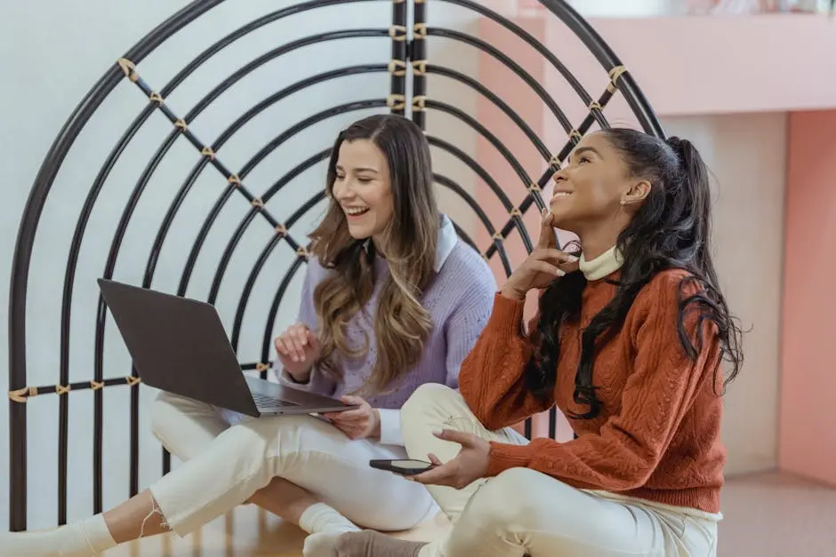 Two diverse women joyfully working together on a laptop indoors, embodying teamwork and digital connectivity.