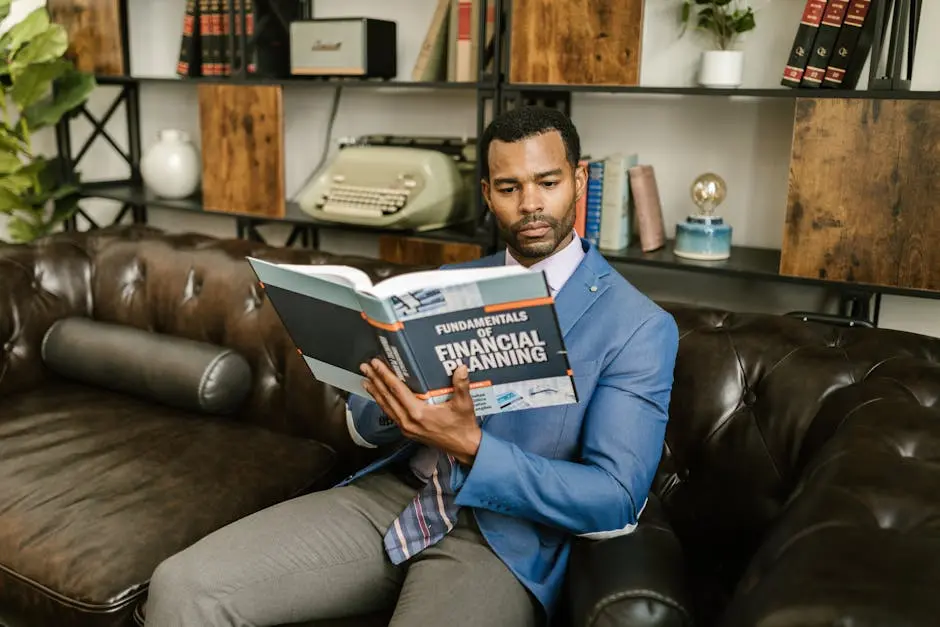 A man in a blue blazer sits on a leather couch reading a financial planning book.