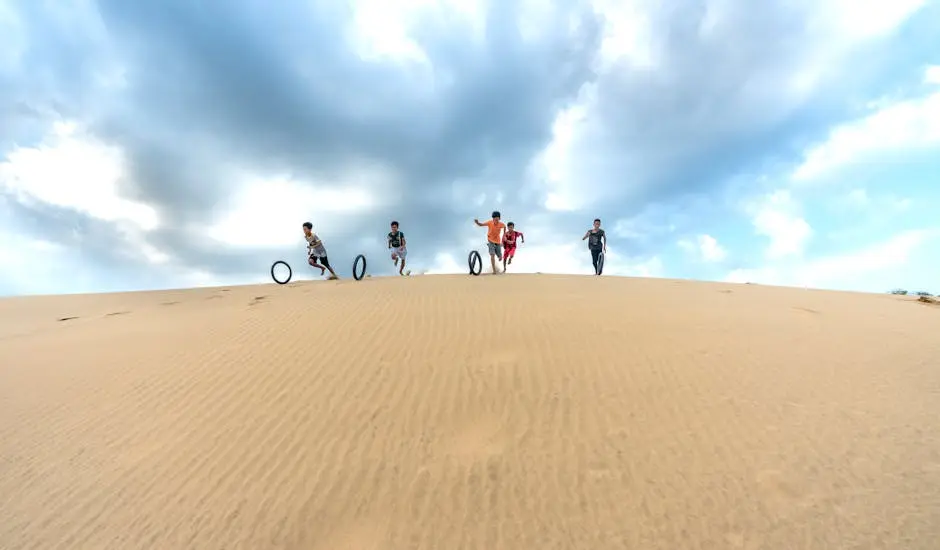 Five children running down sand dunes while playing with tires under a cloudy sky.