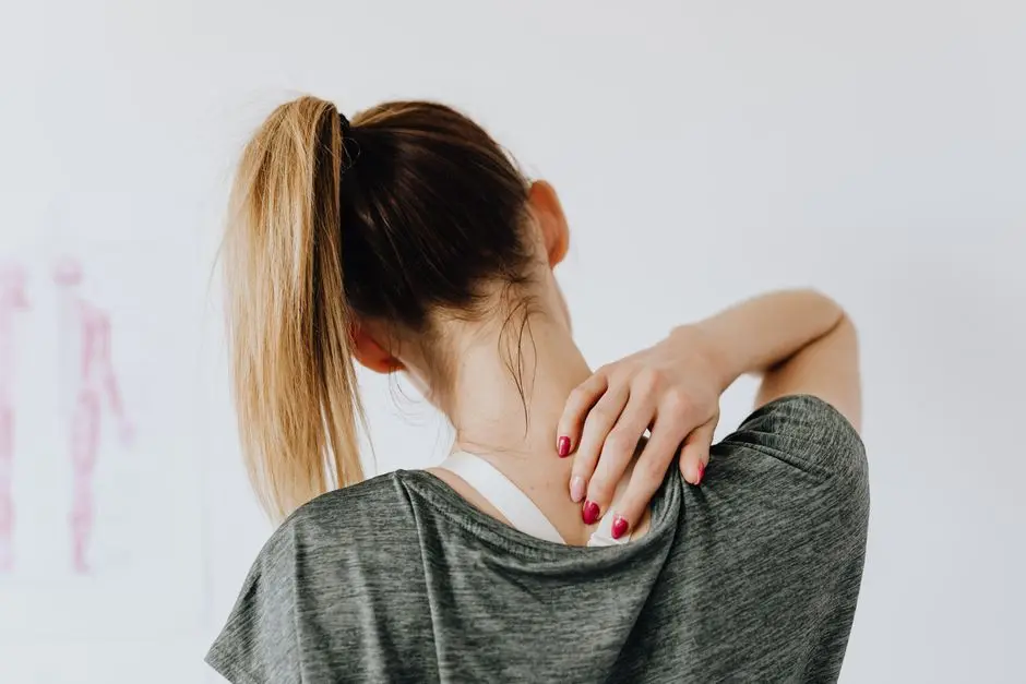 Back view of slim blond female in casual wear with manicure indicating pain spot on back while sitting in front of white wall with paper drawing in clinic