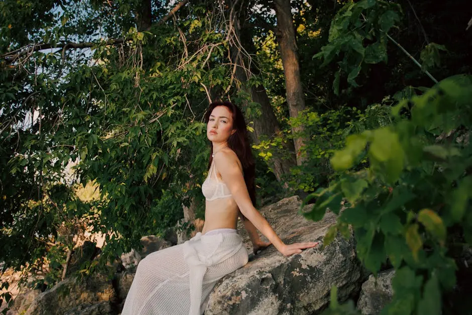 Woman poses outdoors in white attire sitting on a stone ledge surrounded by greenery.