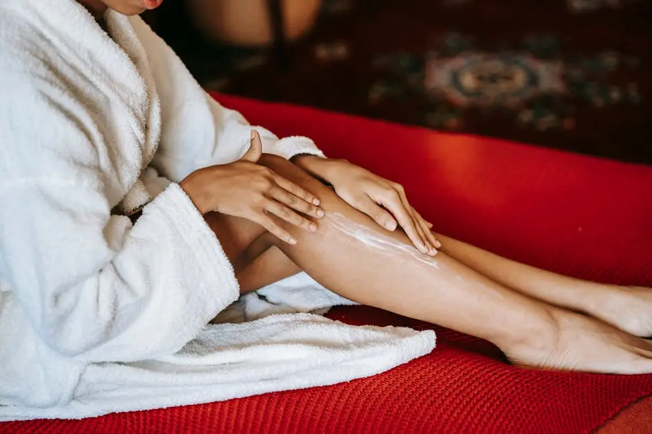 A woman in a bathrobe applies moisturizer to her legs at home, symbolizing relaxation and skincare.