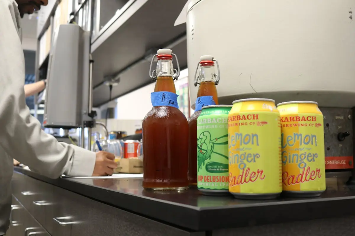 A variety of craft beer bottles and cans on a counter with a man writing in the background.