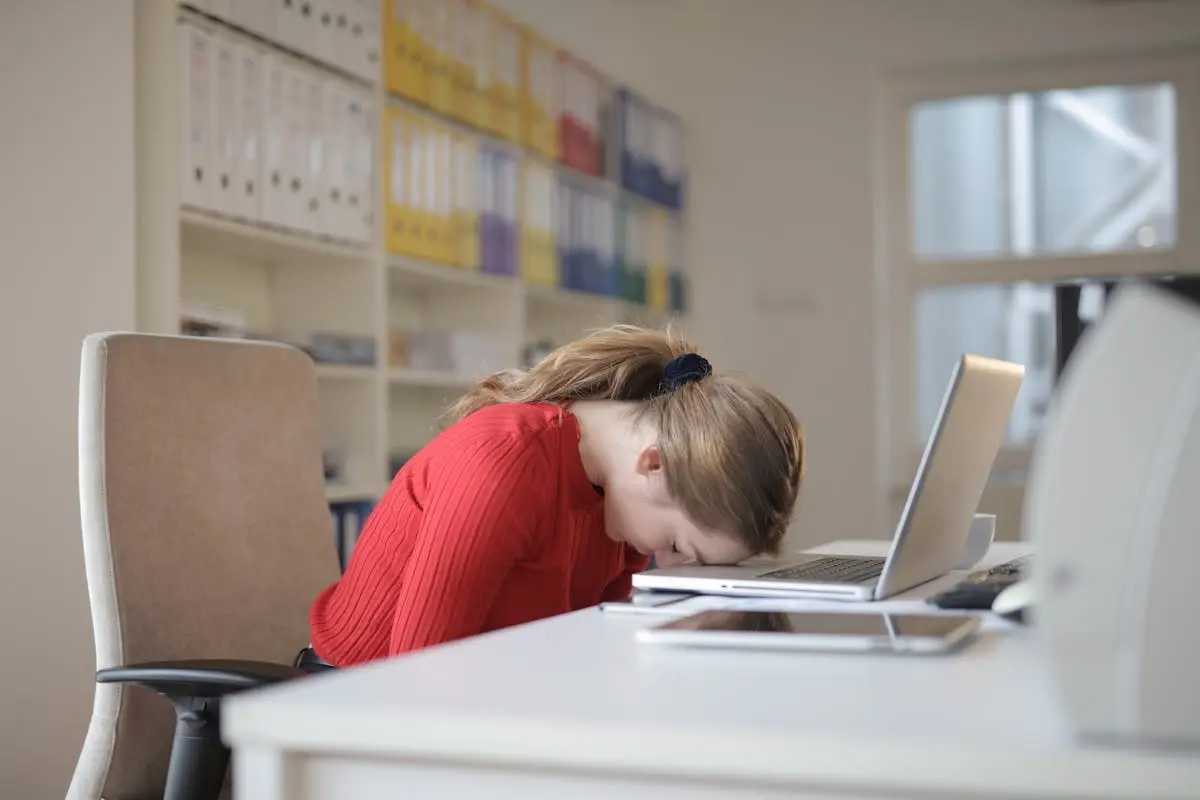 A tired woman in a red sweater leans her head on a desk with a laptop, symbolizing workplace fatigue. Career Change
