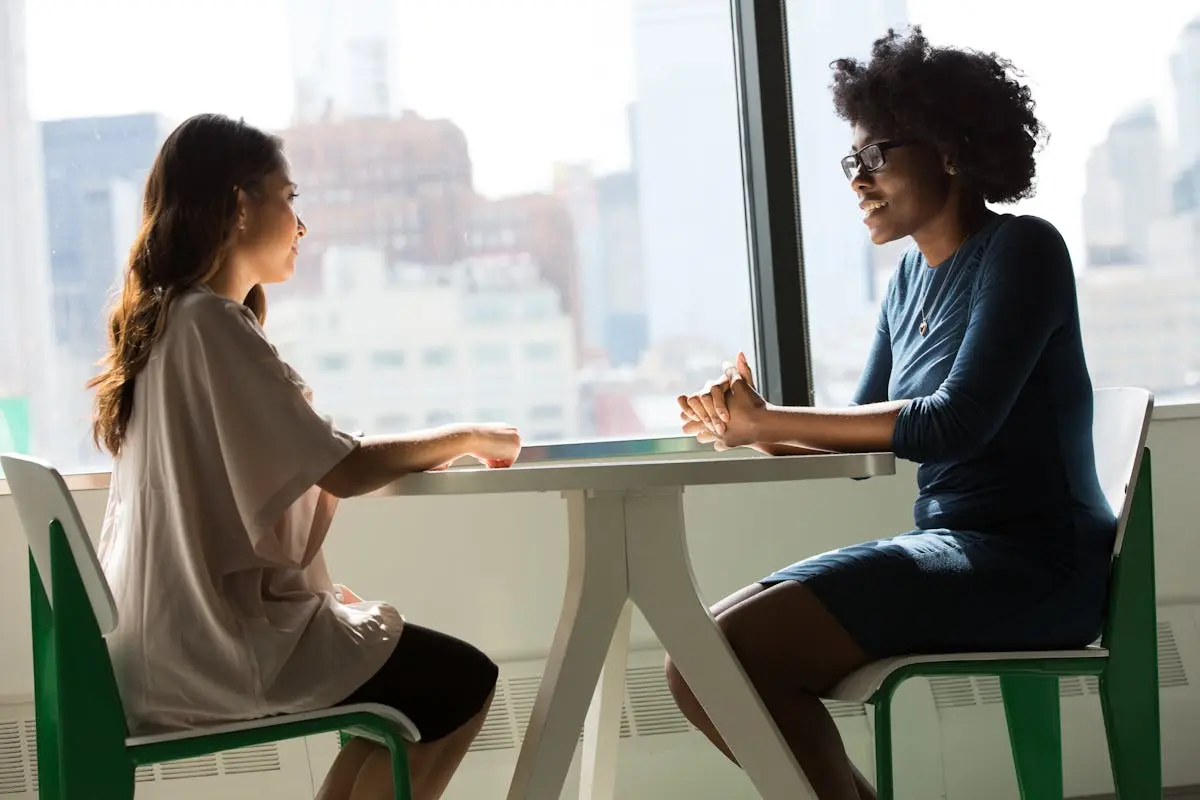 Two women small business consulting sitting and talking at a table with a city view from the window.