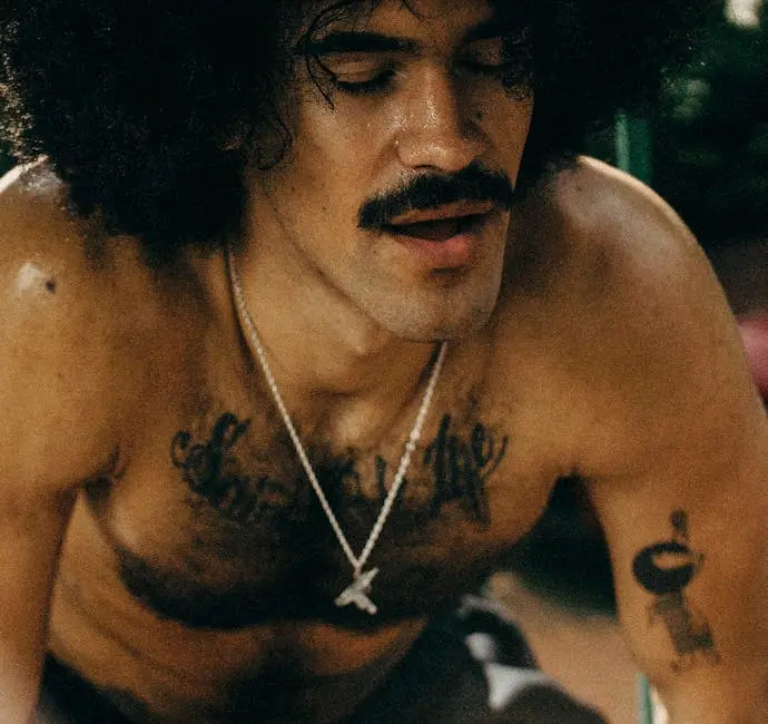 Close-up of a shirtless man with a mustache and afro, showcasing tattoos and jewelry.
