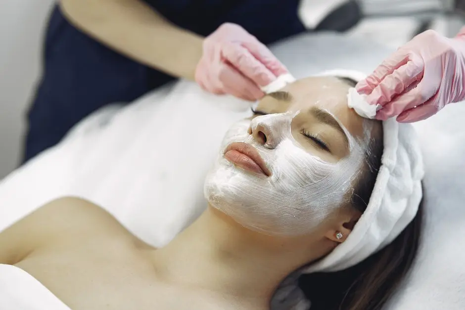 From above anonymous medical specialist in uniform and gloves wiping cream mask off face of relaxed woman during skincare procedure in modern studio