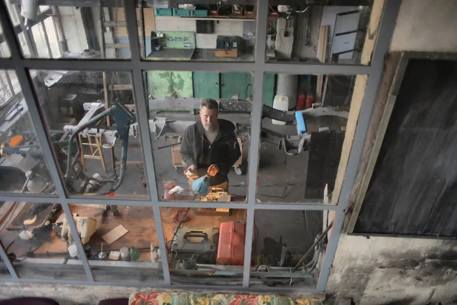 A man focuses on his work in a cluttered industrial workshop, viewed through a window.