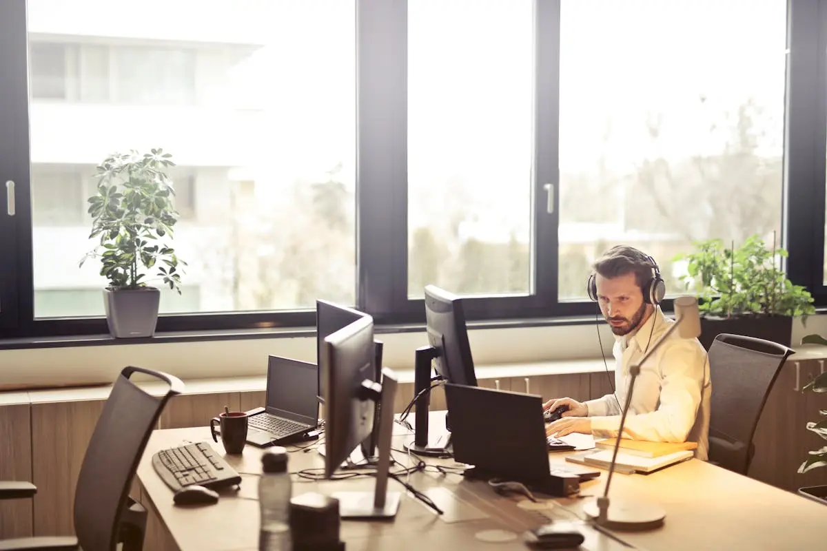 A businessman sits at a desk using multiple computers and a headset in a well-lit modern office.