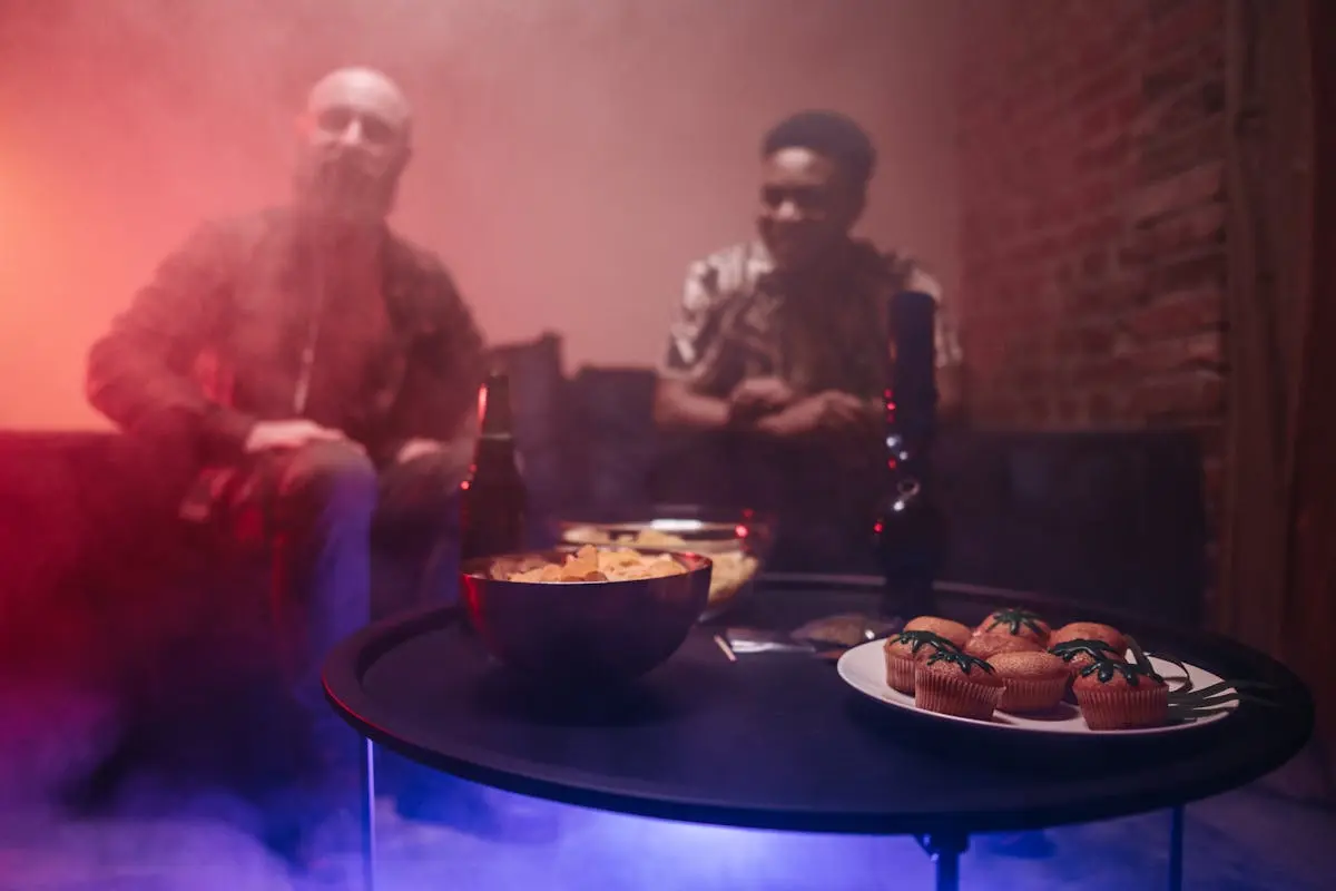 Two people in a dimly lit room with smoke, enjoying drinks and snacks on a table.