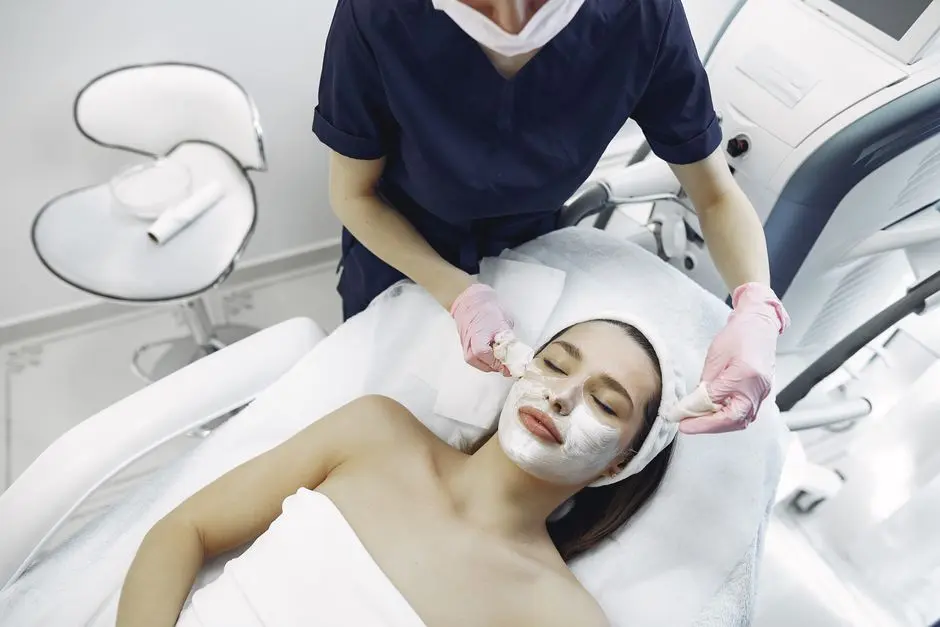 From above of dermatologist in medical coat and latex gloves doing face cleansing and wiping mask from face of female lying in towel in modern salon