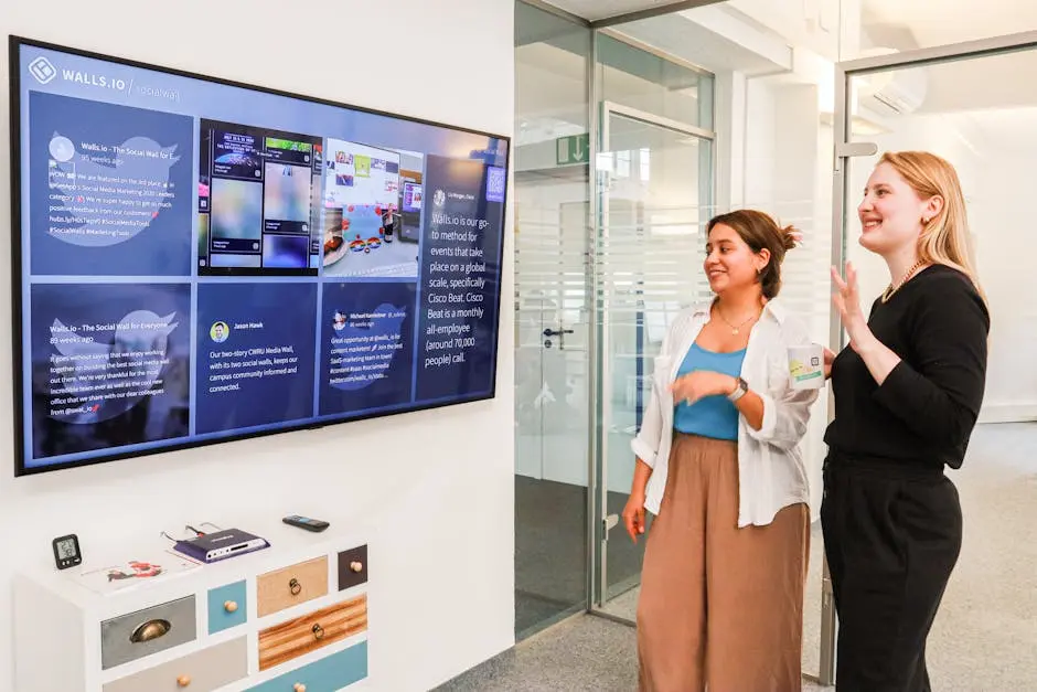 Two women in an office environment interact with a large digital display showcasing social media content.