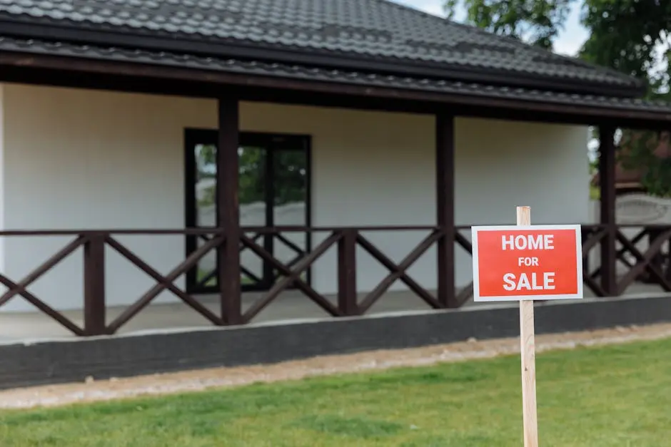 White house with porch and ‘Home for Sale’ sign on a sunny day.