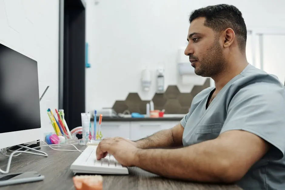 Dentist in scrubs working on a computer at a dental clinic, with focus on patient management.