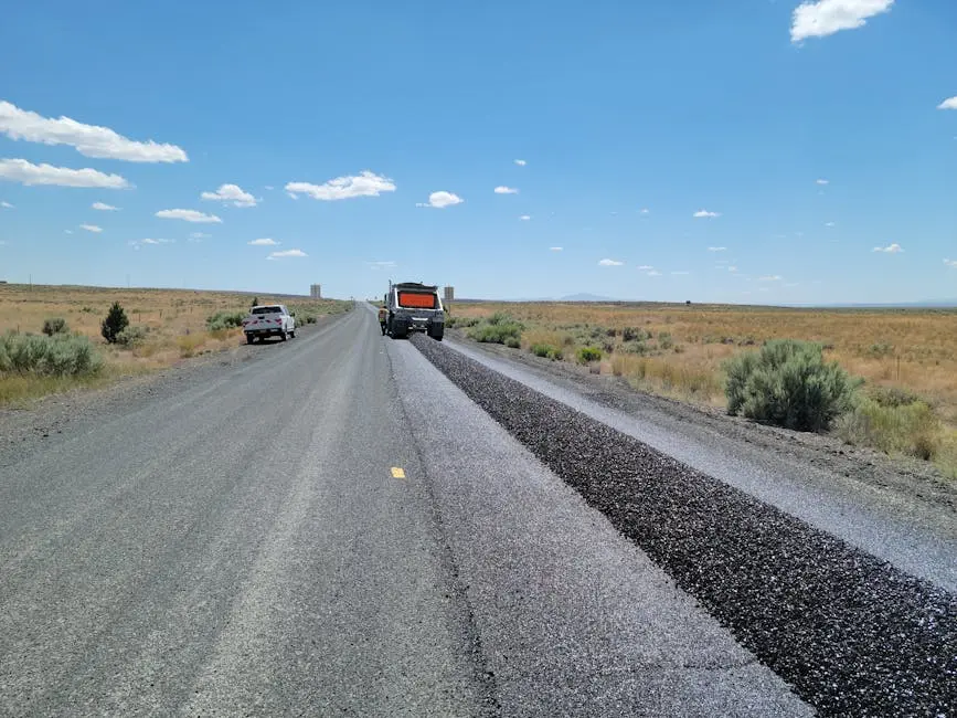 Construction equipment resurfacing a rural highway under a clear blue sky.