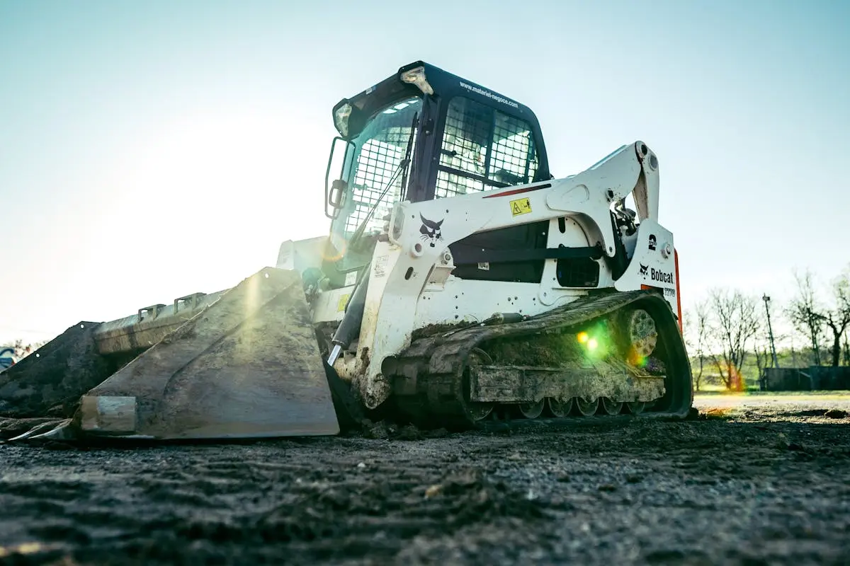 A skid steer loader on a construction site with bright sunlight in the background.