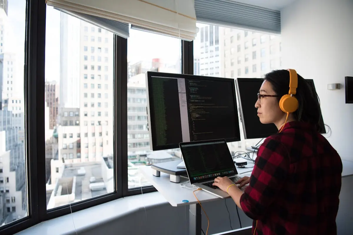 A businesswoman coding at a standing desk with city views through large windows, wearing headphones.