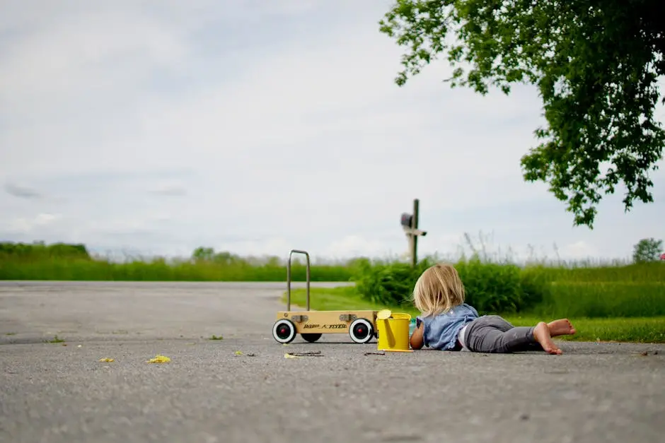 A young child playing on an asphalt road with a toy car and bucket in a rural outdoor setting.