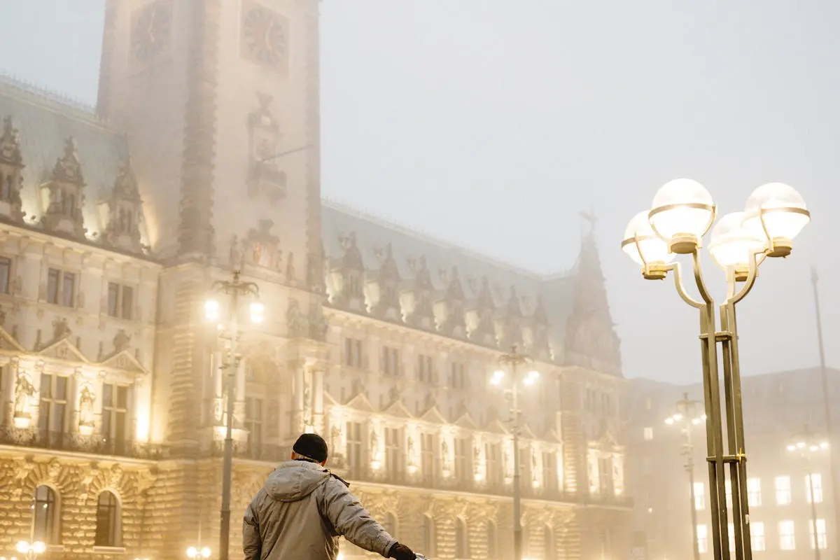 A lone cyclist stands with a bike in front of fog-shrouded Hamburg City Hall at night.