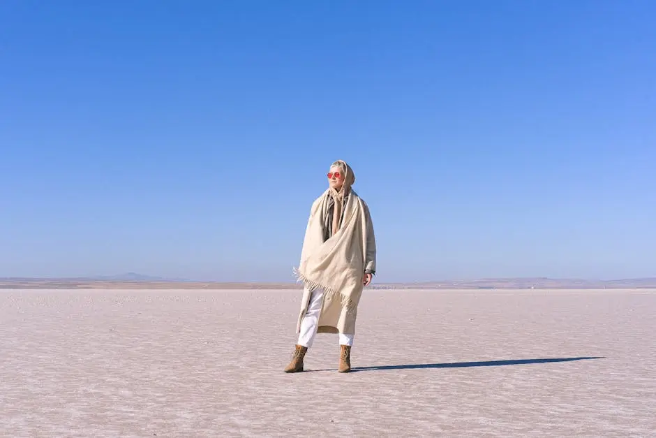 A stylish woman stands in a vast desert landscape, wrapped in a scarf and wearing sunglasses.