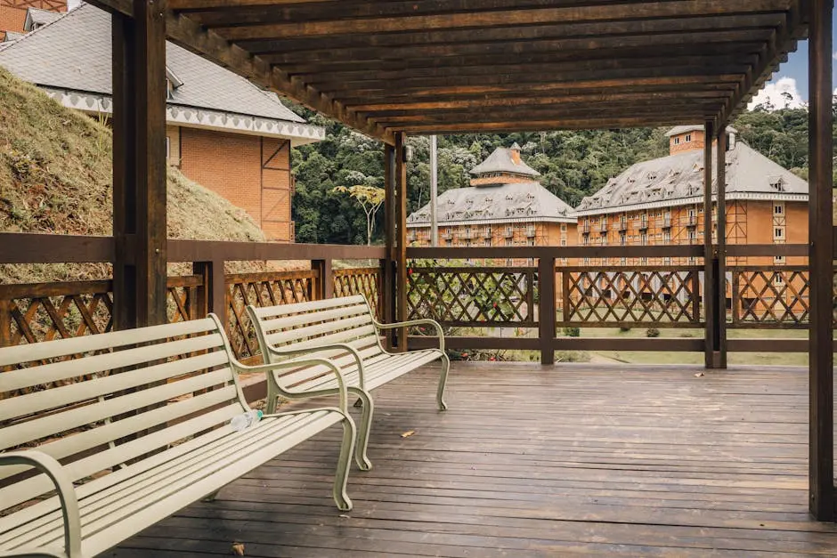 A scenic wooden terrace with benches faces a charming rustic building amidst lush greenery.
