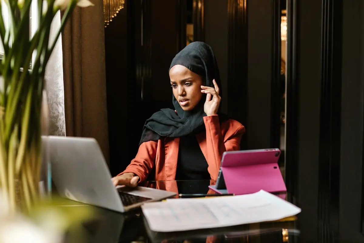 A modern, fashionable woman in a hijab working on her laptop in an elegant indoor setting.