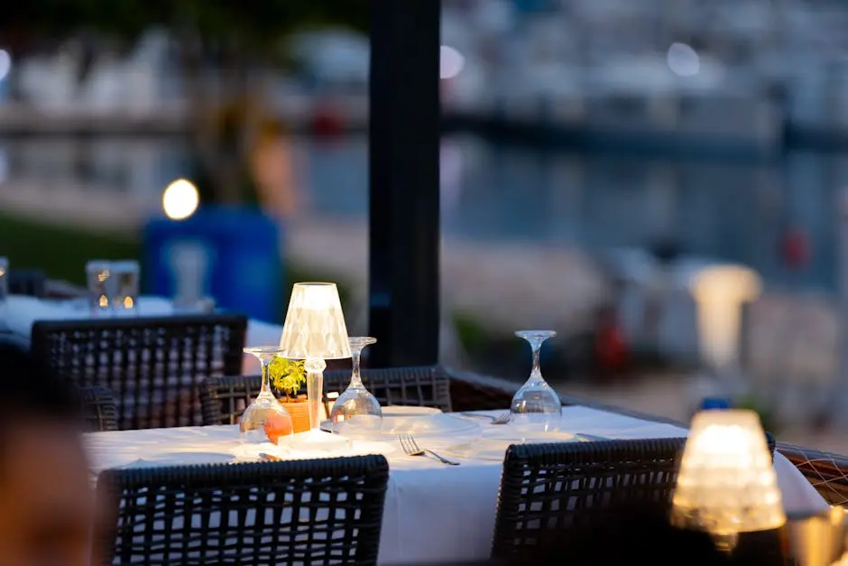 Charming outdoor restaurant setup with lamps and wine glasses by a marina at twilight.