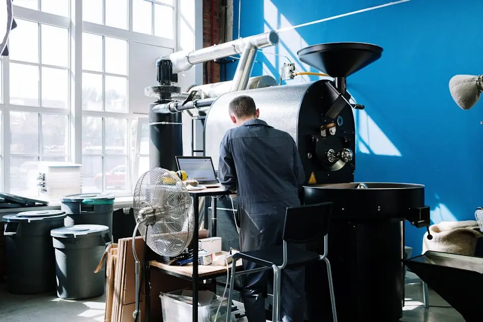 A professional in coveralls operates a coffee roaster in a sunlit workshop.