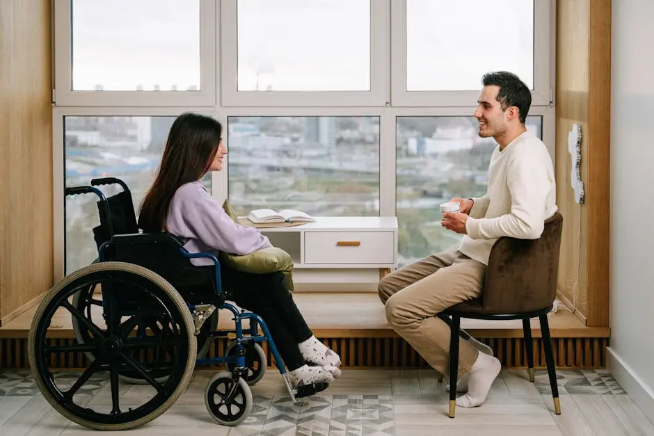 Two adults engaging in a friendly conversation by the window, one in a wheelchair.